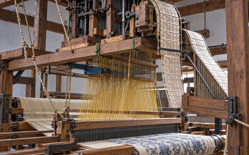A wooden loom with a Jacquard attachment above it: hanging punched cards at the side, harness cords reaching down to the warp, and blue patterned cloth in front.