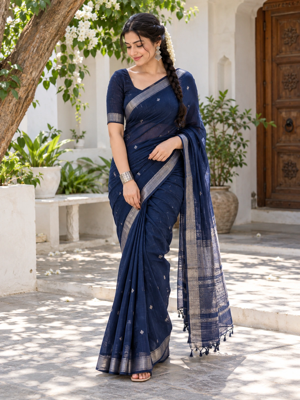 A woman in a navy saree looking down and lightly holding her front pleats in a white courtyard