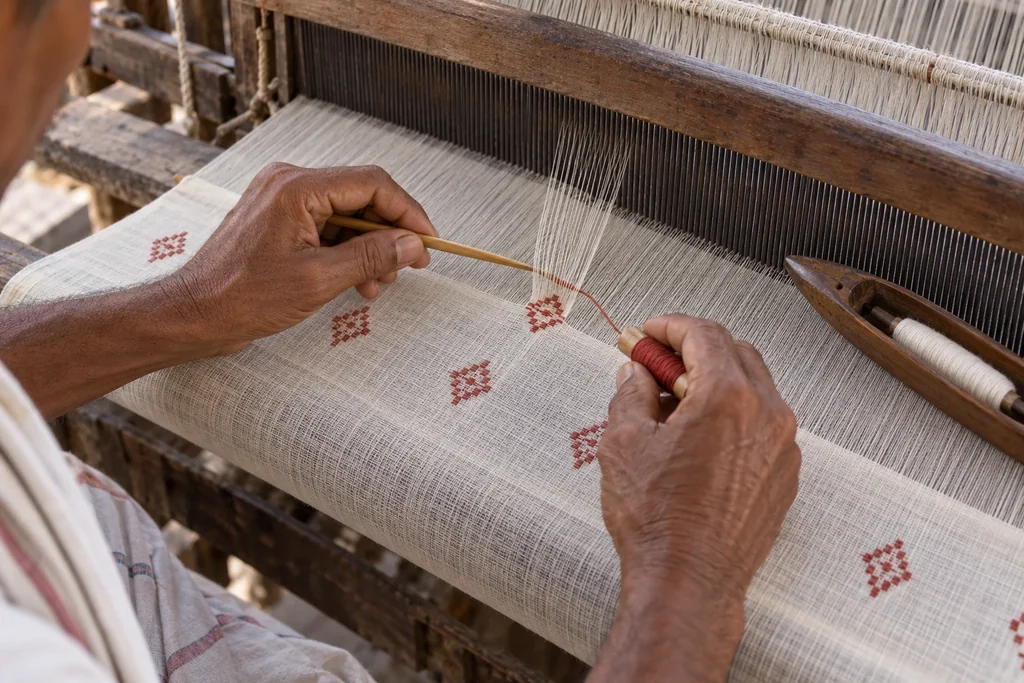 Hands at the edge of fine white cloth on a loom, laying a red pattern thread into a small motif with a slender bamboo tool.